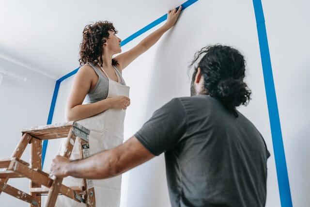 A person standing on a ladder painting a wall while another person holds the ladder